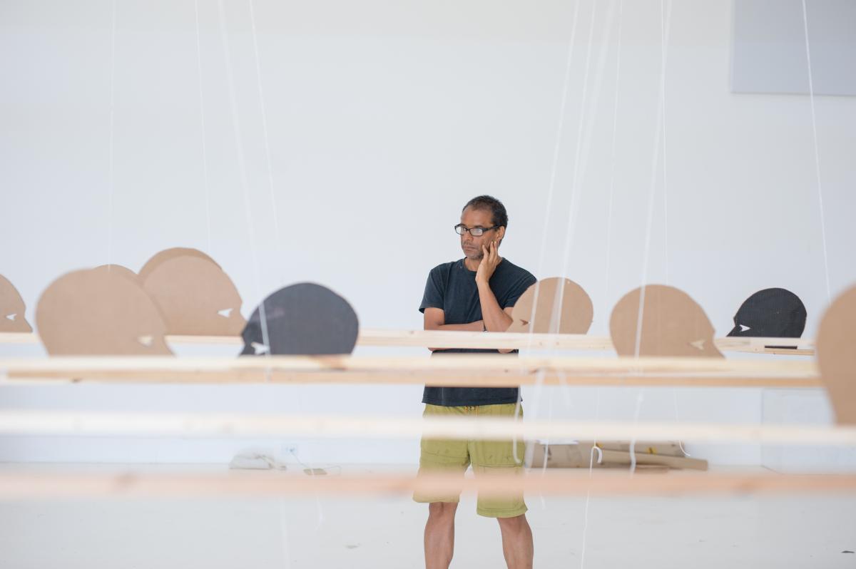 A man stands in the center of a white room with a series of horizontal wooden sticks hanging in front of him with brown and black cardboard head silhouettes.