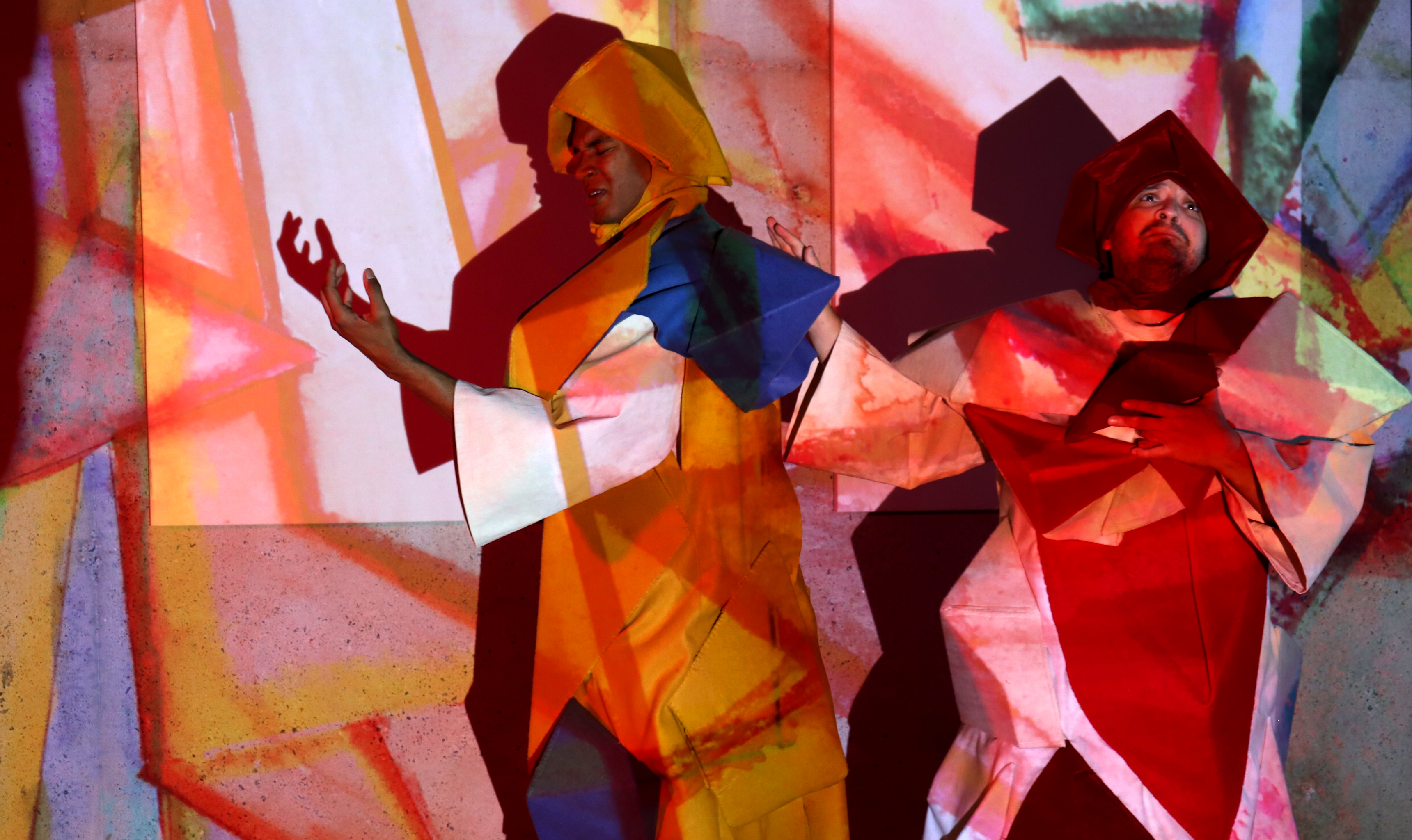 Two people in colorful geometric outfits stand against a vibrant abstract background with projected colors.